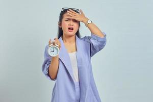 Mujer asiática joven preocupada sosteniendo el despertador con las manos sobre la cabeza aislada sobre fondo blanco