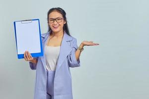 retrato de una joven y alegre mujer de negocios asiática que muestra un portapapeles en blanco y señala el espacio de copia con la palma aislada en un fondo morado foto