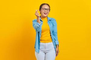 Portrait of smiling young Asian woman student in denim clothes showing korean heart with two fingers crossed isolated on yellow background. People sincere emotions lifestyle concept photo