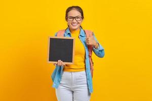Portrait of cheerful young Asian woman student in casual clothes with backpack holding blank chalkboard and showing thumb up isolated on yellow background