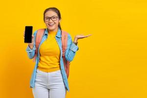 Portrait of cheerful young Asian woman student in casual clothes with backpack holding smartphone and pointing to copy space with palms isolated on yellow background photo