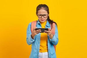 retrato de una joven estudiante asiática seria vestida de forma informal con mochila jugando videojuegos en un teléfono móvil aislada de fondo amarillo. educación en concepto de universidad universitaria foto