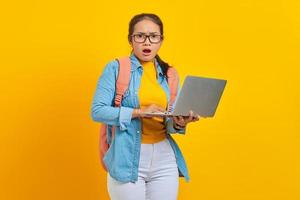 retrato de una joven estudiante asiática sorprendida con ropa informal con mochila usando una laptop y mirando una cámara aislada en un fondo amarillo. educación en concepto de colegio universitario foto