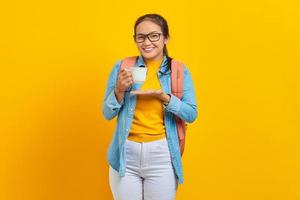 Excited beautiful young Asian woman student in denim clothes with backpack while showing cup of coffee with palms isolated on yellow background photo