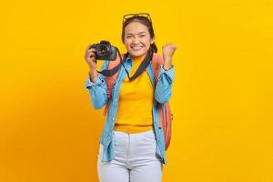 retrato de una joven asiática emocionada con mochila sosteniendo una cámara profesional y celebrando la suerte aislada de fondo amarillo foto