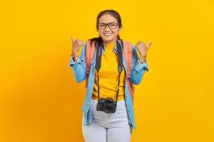Cheerful young traveler Asian woman with backpack and camera in denim clothes while showing thumb up gesture isolated on yellow background. Passenger traveling on weekends. Air flight journey concept
