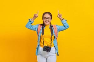 Portrait of cheerful young traveler Asian woman with backpack and camera in denim clothes while pointing at copy space with fingers isolated on yellow background. Air flight journey concept