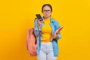 retrato de una joven estudiante asiática triste vestida de denim con mochila usando teléfono móvil y sosteniendo un libro aislado en un fondo amarillo foto