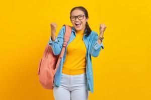 Cheerful young Asian student in denim outfit with backpack standing doing winning gesture celebrating fist saying yes isolated on yellow background. Education in high school university college concept photo