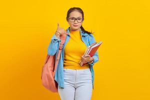 retrato de una joven estudiante asiática sonriente vestida de denim con mochila sosteniendo un cuaderno y haciendo gestos telefónicos como dice llámame aislada en un fondo amarillo foto