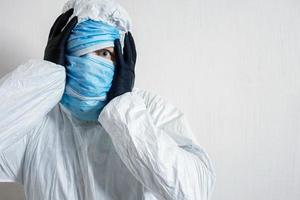 un hombre asustado con un traje protector colgado con máscaras médicas representa el horror contra una pared blanca. los horrores de la epidemia, el peligro del coronavirus foto