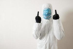 a man in a protective suit hung with medical masks posing against a wall background showing various gestures with his fingers photo