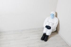 un hombre condenado con un traje protector y muchas máscaras médicas en la cara se sienta en la esquina de la habitación. esconde toda su cara para protegerse del coronavirus. foto
