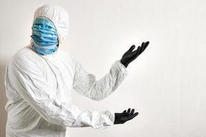 man in a protective suit hung with medical masks posing against wall background showing various gestures with his fingers. the scientist gestures to the wall where the text can be placed photo