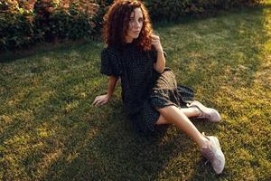 adorable curly red-haired girl with freckles in a dress enjoys good weather sitting on the lawn photo