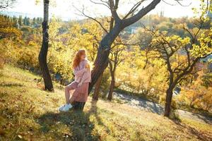 retratos de una encantadora chica pelirroja con una cara linda. chica posando en el parque de otoño con un suéter y una falda de color coral. en manos de una niña una hoja amarilla foto