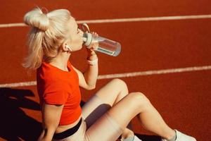 joven y bella rubia se sienta en una pista de jogging con una botella de agua en las manos foto