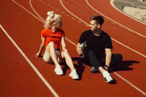 joven hermosa rubia y un chico están sentados en una pista de jogging con una botella de agua y charlando foto