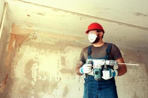 a builder with a perforator poses against the background of a wall with empty space from text The builder is dressed in a protective suit and helmet photo