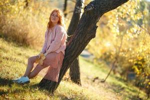 retratos de una encantadora chica pelirroja con una cara linda. chica posando en el parque de otoño con un suéter y una falda de color coral. en manos de una niña una hoja amarilla foto