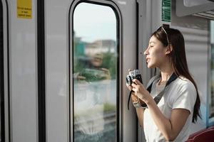 una hermosa turista asiática con cámara en la cabina de pasajeros, viajando en tren aéreo, tomando fotos instantáneas cuando se transporta en vista urbana, estilo de vida de la ciudad en tren, feliz viaje de vacaciones.