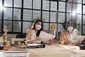 Two female coworkers team with face mask working in new normal office. COVID-19 protection by cleared partition, business workplace office, social distancing for pandemic health, disease prevention. photo