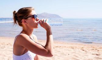 bella mujer con gafas de sol bebiendo agua en la playa tropical foto