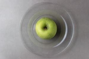 Juicy green whole apple on a plate of transparent glass on a gray background photo