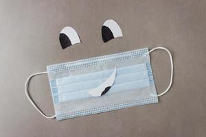 Blue disposable medical mask with a drawn face - smile and eyes on a gray background. The concept of medicine, safety, health, protection from virus, infection photo