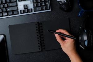 top view hand writing on notebook working on desk with keyboard headphone on black table background photo