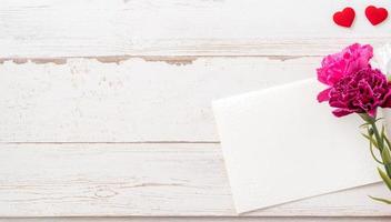 fotografía conceptual del día de las madres de mayo: hermosos claveles y forma de corazón con una tarjeta blanca vacía aislada en una mesa de madera brillante, espacio para copiar, plano, vista superior, maqueta foto