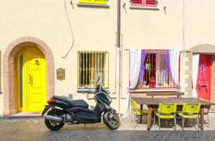 Rimini, Italy, September 19, 2018 table with chairs and motorcycle bike scooter parked on cobblestone street near buildings with colorful wall, door and windows in historical city centre