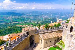 Republic San Marino, September 18, 2018 Aerial top view landscape with valley, green hills, fields, villages of suburban district, blue sky background and stone fortress wall and tower with merlons photo