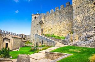 Republic San Marino, September 18, 2018 Courtyard with green grass lawn of Prima Torre Guaita first medieval tower with stone brick fortress wall with merlons on Mount Titano rock with blue sky photo