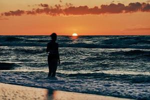 Female silhouette in blue sea waves at summer sunset, half sun below horizon, beachfront holiday photo