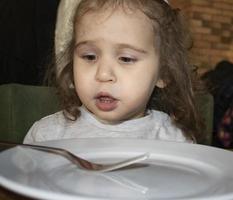 retrato de una linda niña sorprendida sentada en la mesa. mirando el plato vacío. la niña quiere comer. foto