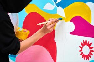 artista con capucha negra pintando un cuadro colorido con pincel sobre lienzo blanco en el festival de arte foto