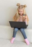 Happy cute caucasian little girl holding credit card buying online sitting on the couch photo