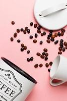 Coffee beans out of the coffee pot with the pot lid next to it and a white cup on a pink background.