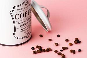 Coffee beans out of coffee pot with pot lid next to it on pink background.