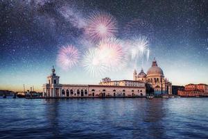 Night illumination of buildings and boats near the water. Photo greeting card. Colorful fireworks on the black sky background