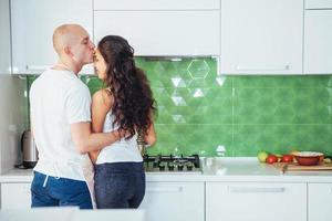 hermosa pareja joven está hablando, mirando a la cámara y sonriendo mientras cocina en la cocina. foto