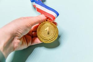 Simply flat lay design hand holding winner or champion gold trophy medal isolated on blue colorful background. Victory first place of competition. Winning or success concept. Top view copy space. photo