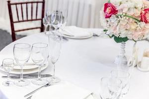 Fancy table set for dinner with flower composition in restaurant, luxury interior background. Wedding elegant banquet decoration and items for food arranged by catering service on white table. photo