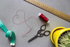 Sewing accessories scissors, measuring tape, ruler, fabric. In center of composition - red thread, laid out in the shape of a heart, with a needle at the end. With cactus for home furnishings photo