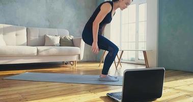 mooie slanke vrouw in een donker trainingspak gaat op de mat bij de laptop op de vloer zitten om deel te nemen aan online training in slow motion in de kamer video