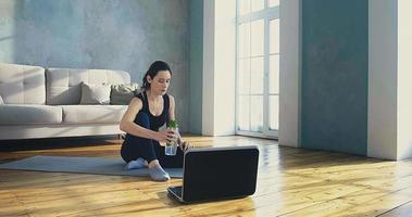 femme sportive brune boit de l'eau reposant sur le sol après l'entraînement et regarde la vidéo sur un ordinateur portable dans une pièce spacieuse au ralenti video
