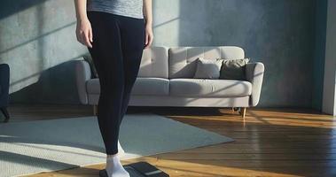 Worried woman in sportswear stands on digital scales becoming happy of slimming down near sofa in living room slow motion video