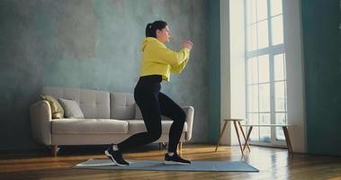 mulher morena de capuz amarelo faz treinamento dinâmico de lunges no tapete perto do sofá na sala de estar em casa video
