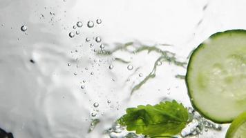 Person throws slices of cucumber lime and mint leaves into clear water on white background macro bottom view slow motion